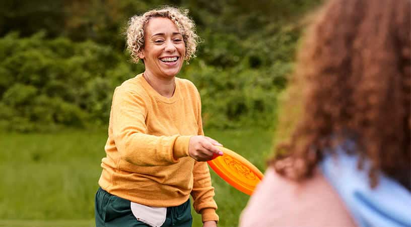 Frau mit Stomabeutel am Bauch und orangenem Pullover spielt draußen Frisbee mit einer anderen Person